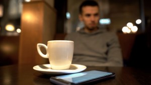 samotny mężczyzna siedzi w kawiarni, a przed na stole stoi filiżanka kawy espresso i leży smartfon