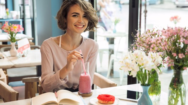 uśmiechnięta kobieta siedzi w kawiarni i pije smoothie, a przed nią leżą książki i pączek na talerzu