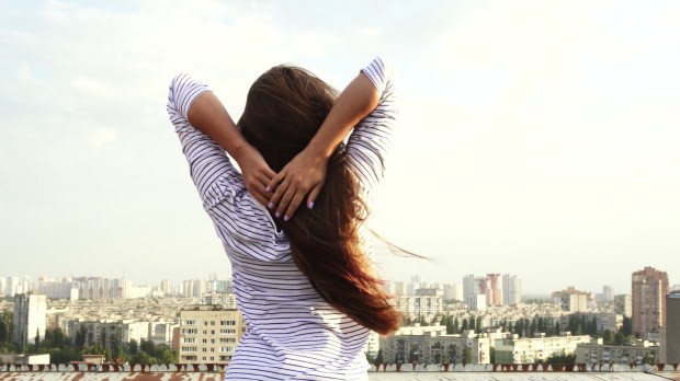 młoda dziewczyna stoi na dachu podziwiając panoramę dużego miast i przytrzymuje rękami swoje długie włosy rozwiewane przez wiatr