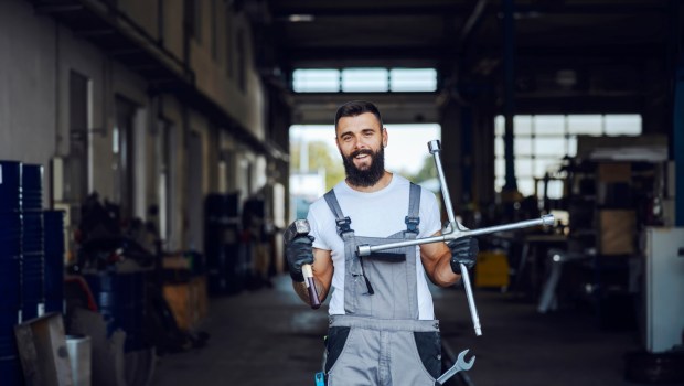 Uśmiechnięty mechanik samochodowy stoi przed warsztatem i trzyma w rękach narzędzie