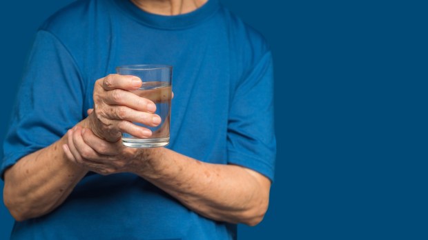 Chory na Parkinsona cżłowiek trzyma z niepewnością szklankę wody, pomagając sobie drugą ręką