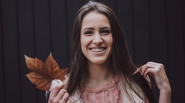 Uśmiechnięta radosna młoda kobieta poprawia włosy i trzyma w ręku jesienny liść